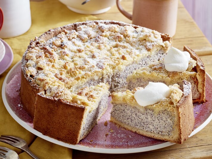 Auf einem Teller ist ein Streusel-Mohn-Käsekuchen serviert. Er ist mit Puderzucker bestreut und wird mit einem Klecks Sahne serviert. | © EAT CLUB / Isabell Triemer