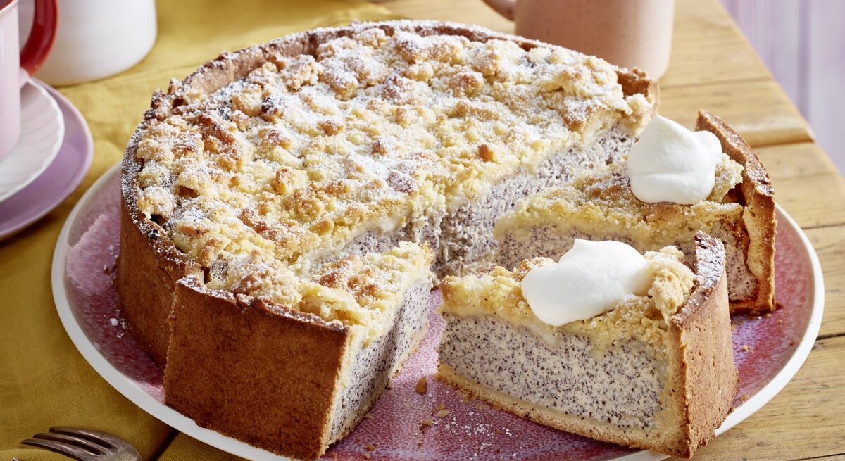 Auf einem Teller ist ein Streusel-Mohn-Käsekuchen serviert. Er ist mit Puderzucker bestreut und wird mit einem Klecks Sahne serviert.