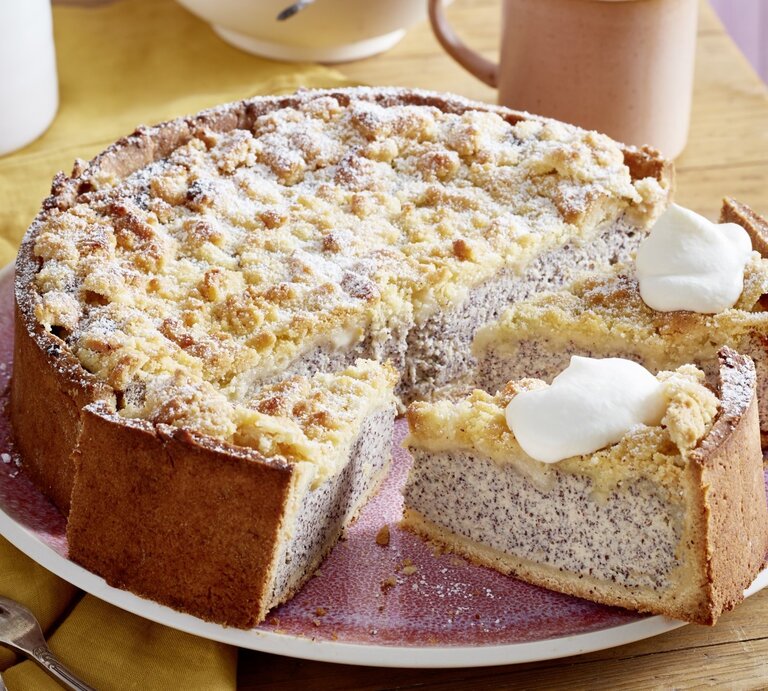 Auf einem Teller ist ein Streusel-Mohn-Käsekuchen serviert. Er ist mit Puderzucker bestreut und wird mit einem Klecks Sahne serviert. | © EAT CLUB / Isabell Triemer