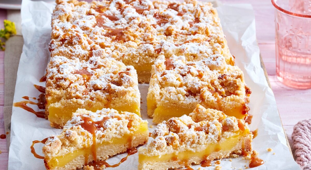 Auf einem Backblech liegt ein Streusel-Puddingkuchen. Er ist bestreut mit Puderzucker und mit Karamellsoße überzogen. Man erkennt die cremige Pudding-Füllung. 