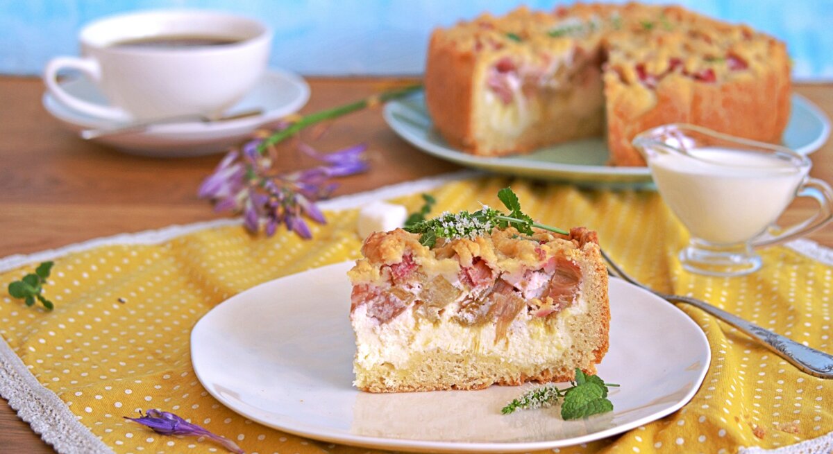 Ein Stück vom Rhabarberkuchen auf einem weißen Teller und einer gelb-blauen Tischdecke. Dahinter eine Tasse Kaffee und der angeschnittene Kuchen. 