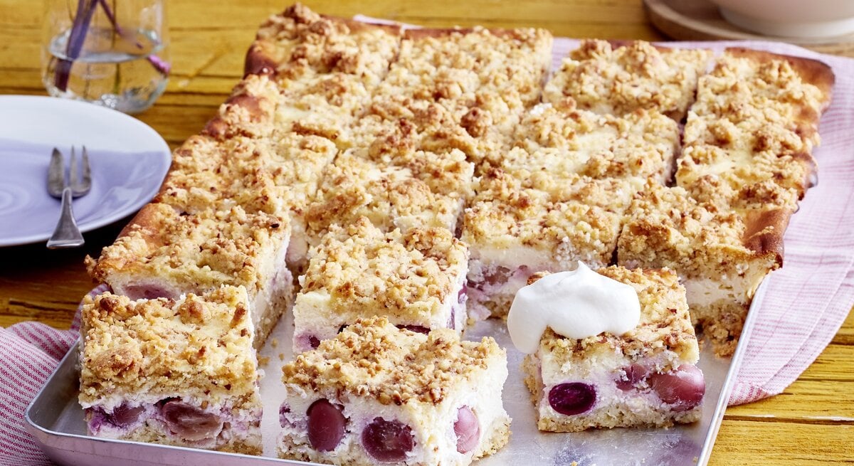 Blechkuchen als Käsekuchen mit Streuseln und Traubenfüllung mit einer Haube aus Sahne auf einem rosaroten Tuch, daneben Blumen 