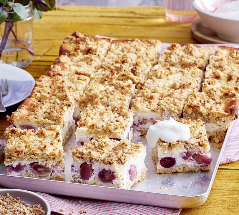 Blechkuchen als Käsekuchen mit Streuseln und Traubenfüllung mit einer Haube aus Sahne auf einem rosaroten Tuch, daneben Blumen  | © EAT CLUB/Isabell Triemer