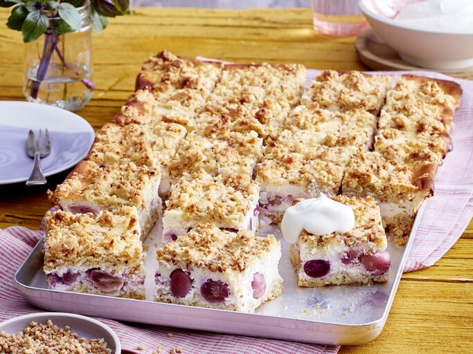 Blechkuchen als Käsekuchen mit Streuseln und Traubenfüllung mit einer Haube aus Sahne auf einem rosaroten Tuch, daneben Blumen  | © EAT CLUB/Isabell Triemer