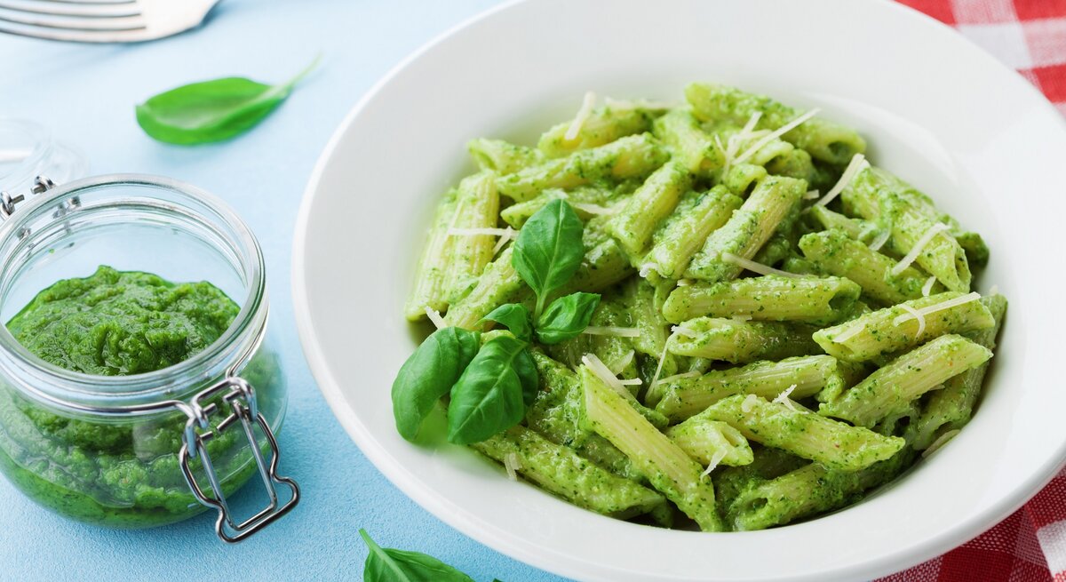Ein Teller Penne mit grünem Miso-Pesto, Basilikum und Streukäse, daneben ein Glas mit dem Pesto sowie eine Gabel und eine rot-weiß-karierte Serviette.