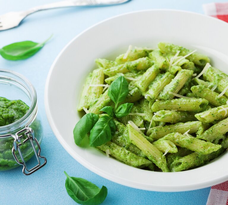Ein Teller Penne mit grünem Miso-Pesto, Basilikum und Streukäse, daneben ein Glas mit dem Pesto sowie eine Gabel und eine rot-weiß-karierte Serviette. | © Adobe Stock/ juliasudnitskaya