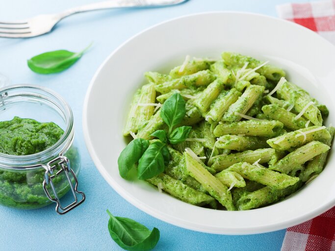 Ein Teller Penne mit grünem Miso-Pesto, Basilikum und Streukäse, daneben ein Glas mit dem Pesto sowie eine Gabel und eine rot-weiß-karierte Serviette. | © Adobe Stock/ juliasudnitskaya