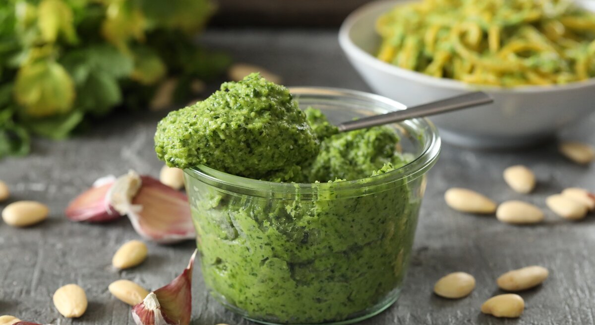 Frontal: eine Glas selbstgemachtes Mandel-Petersilien-Pesto steht auf einem dunklen Untergrund aus Holt. Drum herum liegen Mandeln ohne Haut und ungeschälte Knoblauczehen.