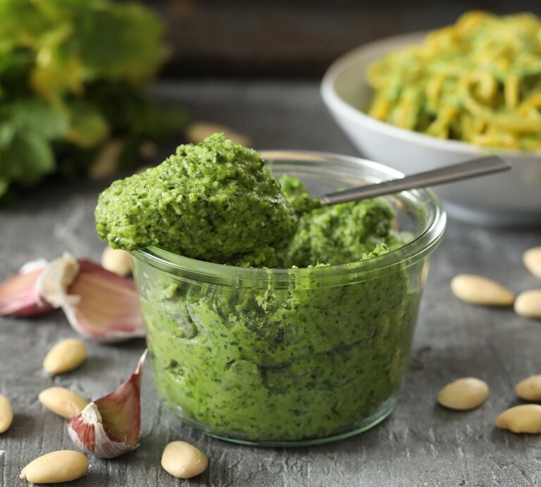Frontal: eine Glas selbstgemachtes Mandel-Petersilien-Pesto steht auf einem dunklen Untergrund aus Holt. Drum herum liegen Mandeln ohne Haut und ungeschälte Knoblauczehen. | © Shutterstock / Joanna Grabowska-Kowalska