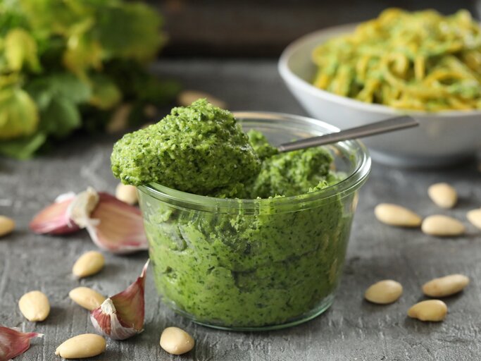 Frontal: eine Glas selbstgemachtes Mandel-Petersilien-Pesto steht auf einem dunklen Untergrund aus Holt. Drum herum liegen Mandeln ohne Haut und ungeschälte Knoblauczehen. | © Shutterstock / Joanna Grabowska-Kowalska