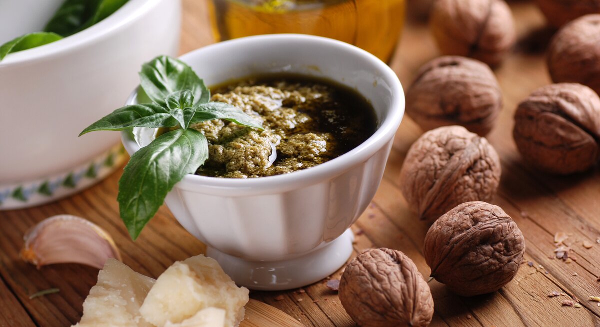 Frontalsicht: Auf einem Untergrund aus Holz steht ein weißes Schälchen selbst gemachtes Walnuss-Pesto mit einem Basilikumblatt als Deko. Drumherum liegen frische Walnüsse, geriebener Parmesan. Im Hintergrund ist eine Glasflasche mit Olivenöl.