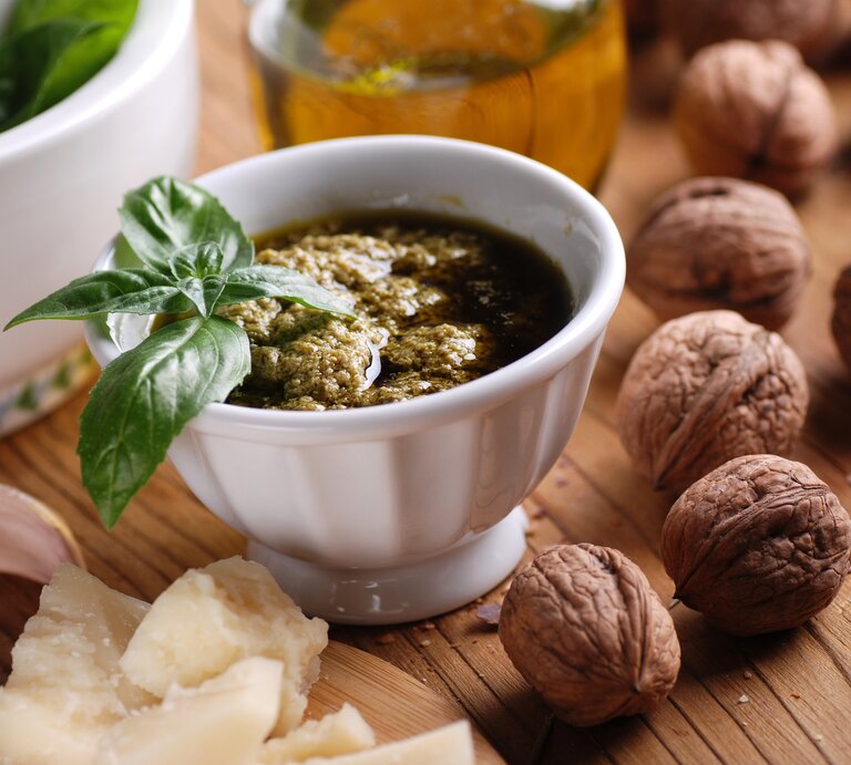 Frontalsicht: Auf einem Untergrund aus Holz steht ein weißes Schälchen selbst gemachtes Walnuss-Pesto mit einem Basilikumblatt als Deko. Drumherum liegen frische Walnüsse, geriebener Parmesan. Im Hintergrund ist eine Glasflasche mit Olivenöl. | © Shutterstock / 	al62