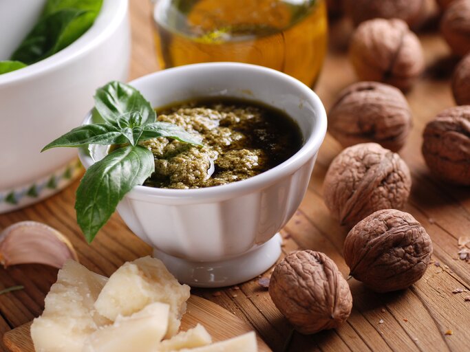 Frontalsicht: Auf einem Untergrund aus Holz steht ein weißes Schälchen selbst gemachtes Walnuss-Pesto mit einem Basilikumblatt als Deko. Drumherum liegen frische Walnüsse, geriebener Parmesan. Im Hintergrund ist eine Glasflasche mit Olivenöl. | © Shutterstock / 	al62