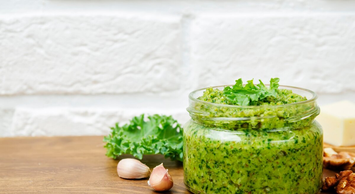 Ein Glas Grünkohlpesto, daneben Knoblauchzehen, Walnüsse und frischer Grünkohl.