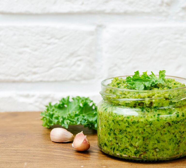 Ein Glas Grünkohlpesto, daneben Knoblauchzehen, Walnüsse und frischer Grünkohl. | © Adobe Stock/ nata_vkusidey 