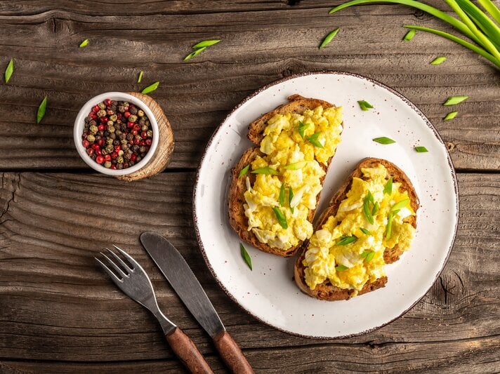 Rührei auf Brot, serviert auf weißem Teller. Dieser steht auf Holztisch. Daneben liegen Frühlingszwiebeln und Pfefferkörner.  | © Getty Images/Koval Nadiya