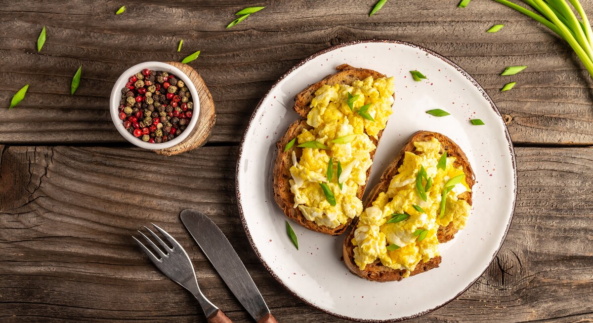 Rührei auf Brot, serviert auf weißem Teller. Dieser steht auf Holztisch. Daneben liegen Frühlingszwiebeln und Pfefferkörner. 