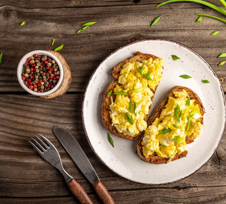 Rührei auf Brot, serviert auf weißem Teller. Dieser steht auf Holztisch. Daneben liegen Frühlingszwiebeln und Pfefferkörner.  | © Getty Images/Koval Nadiya