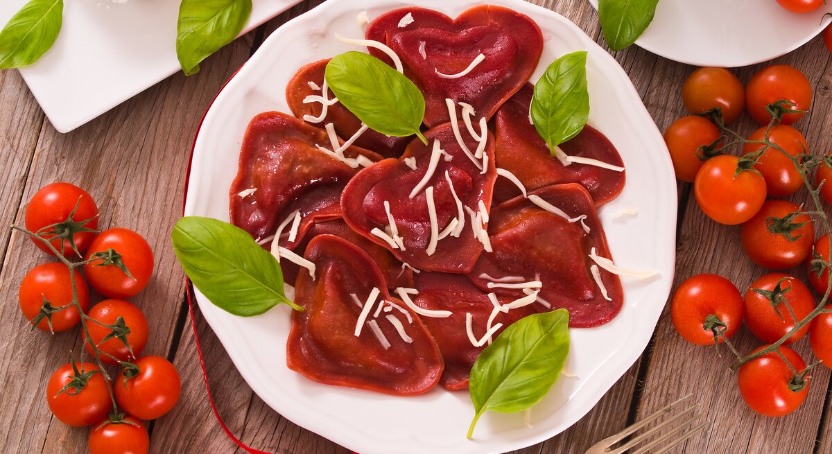 Einige rote Herz-Ravioli aus Rote-Bete-Saft und Tomatenmark mit Hackfleischfüllung liegen auf einem weißen Teller. Die Nudeln sind mit Streukäse und Basilikum angerichtet.