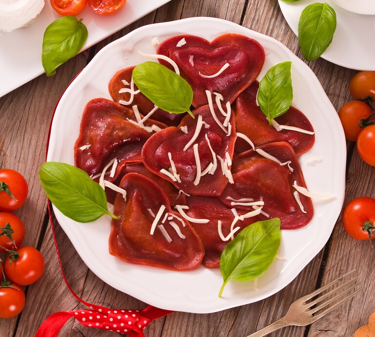 Einige rote Herz-Ravioli aus Rote-Bete-Saft und Tomatenmark mit Hackfleischfüllung liegen auf einem weißen Teller. Die Nudeln sind mit Streukäse und Basilikum angerichtet. | © Getty Images / milla1974
