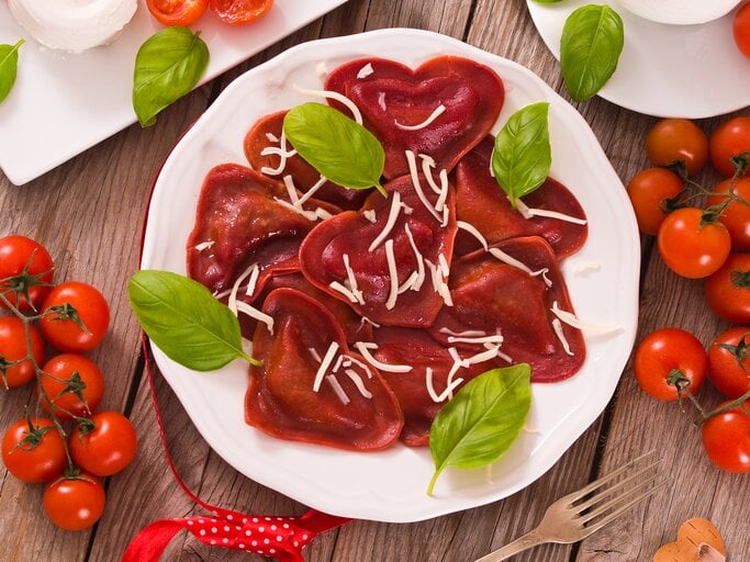 Einige rote Herz-Ravioli aus Rote-Bete-Saft und Tomatenmark mit Hackfleischfüllung liegen auf einem weißen Teller. Die Nudeln sind mit Streukäse und Basilikum angerichtet. | © Getty Images / milla1974