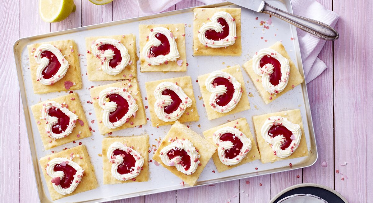 Auf einem Backblech liegen einige Stücken des Zitronen-Herz-Kuchen. Sie sind dekoriert mit herförmiger Mascarpone-creme und Erdbeerkonfitüre. 