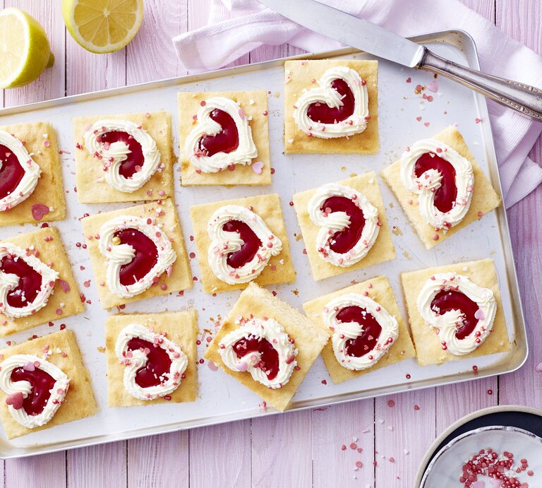 Auf einem Backblech liegen einige Stücken des Zitronen-Herz-Kuchen. Sie sind dekoriert mit herförmiger Mascarpone-creme und Erdbeerkonfitüre.  | © EAT CLUB/Isabell Triemer