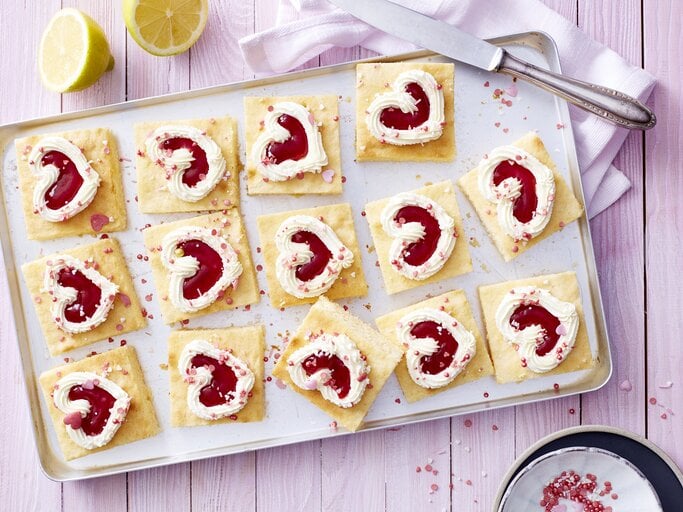 Auf einem Backblech liegen einige Stücken des Zitronen-Herz-Kuchen. Sie sind dekoriert mit herförmiger Mascarpone-creme und Erdbeerkonfitüre.  | © EAT CLUB/Isabell Triemer