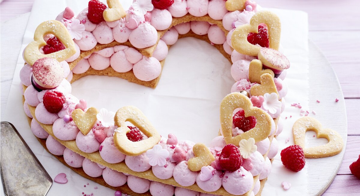 Auf einem Teller liegt ein Herz-Lettercake. Er ist dekoriert mit Zuckerperlen, Keks-Herzen, Himbeeren und rosa Creme. 