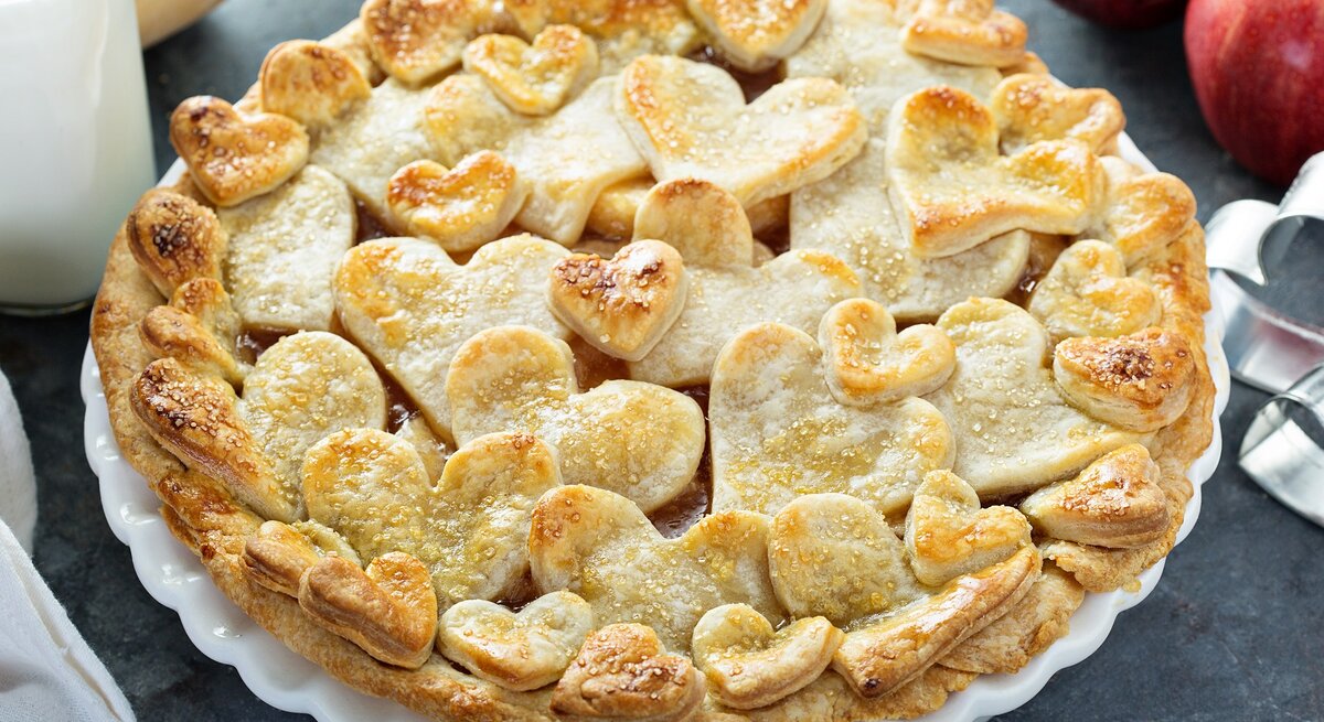 Auf einem Teller ist ein Apfel-Gitterkuchen mit Herzen serviert. Er hat Teigförmige Herzen oben drauf.
