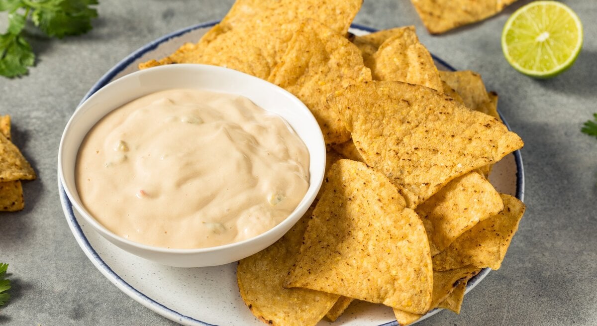 White Queso Dip mit Nachos auf weißem Teller und grauem Tisch. Im Hintergrund Limette, Nachos und Koriander. Draufsicht.