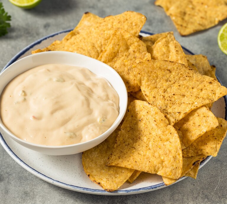 White Queso Dip mit Nachos auf weißem Teller und grauem Tisch. Im Hintergrund Limette, Nachos und Koriander. Draufsicht. | © Getty Images / bhofack2 