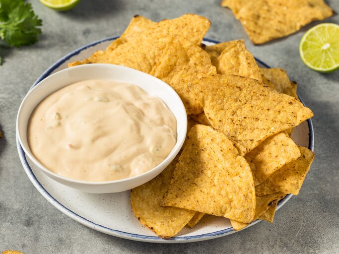White Queso Dip mit Nachos auf weißem Teller und grauem Tisch. Im Hintergrund Limette, Nachos und Koriander. Draufsicht. | © Getty Images / bhofack2 