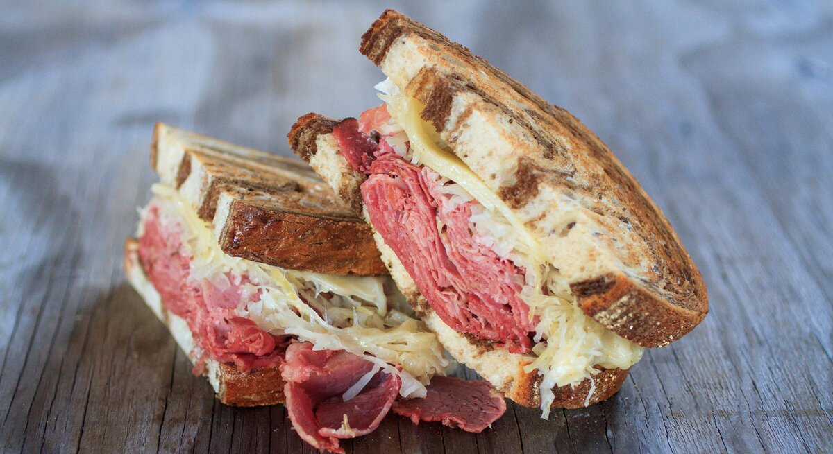 Ein halbiertes Pastrami Sandwich auf einem Holzuntergrund. Alles nah und von der Seite fotografiert.