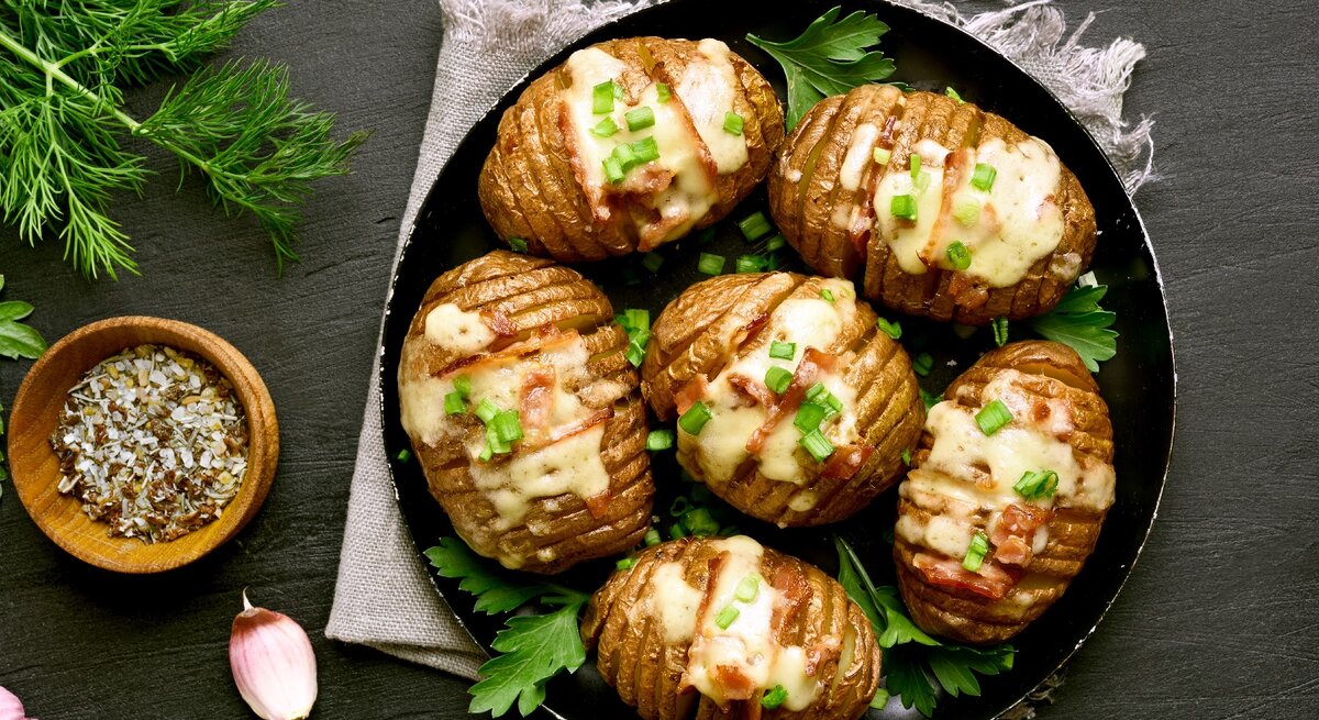 Eine Pfanne mit den Hasselback-Kartoffeln auf einer grauen Decke und einem Schiefer-Untergrund.