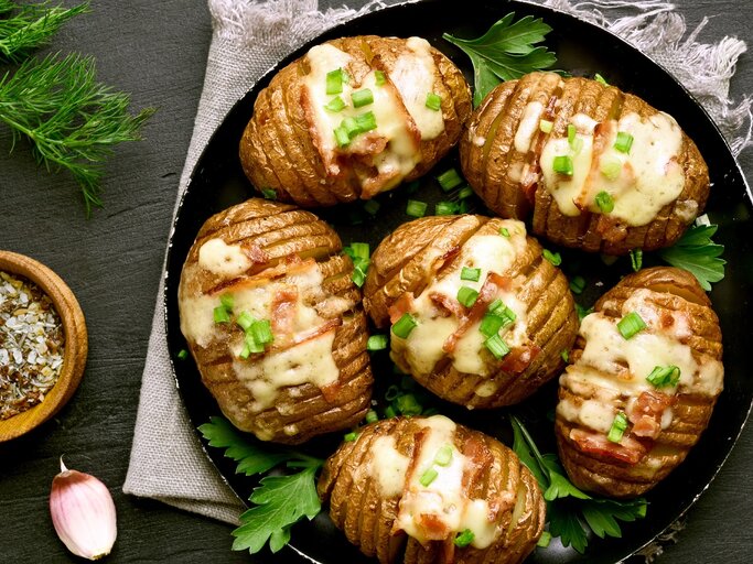 Eine Pfanne mit den Hasselback-Kartoffeln auf einer grauen Decke und einem Schiefer-Untergrund. | © Shutterstock/ Tatiana Volgutova