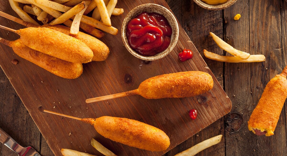 Corn-Dogs mit Pommes auf einem Holzbrett. Daneben kleine Schüssel mit Ketchup und Senf. 