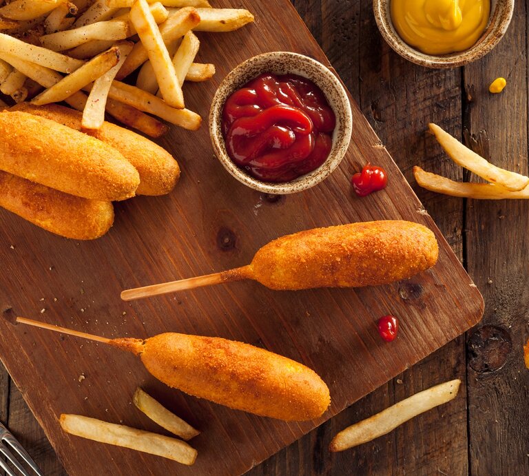 Corn-Dogs mit Pommes auf einem Holzbrett. Daneben kleine Schüssel mit Ketchup und Senf.  | © Getty Images/ bhofack2