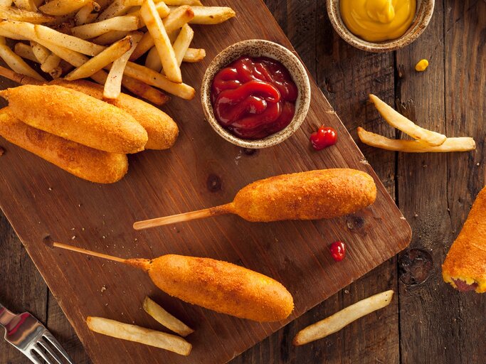 Corn-Dogs mit Pommes auf einem Holzbrett. Daneben kleine Schüssel mit Ketchup und Senf.  | © Getty Images/ bhofack2