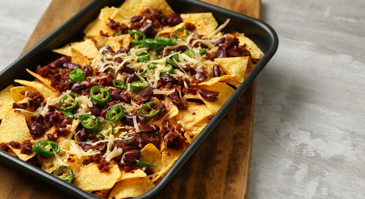 Eine schwarze Ofenform mit überbackenen Nachos mit Hack auf einem Holzbrett.