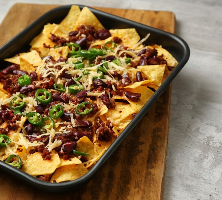 Eine schwarze Ofenform mit überbackenen Nachos mit Hack auf einem Holzbrett. | © AdobeStock/Pixel-Shot