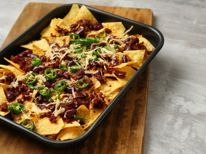 Eine schwarze Ofenform mit überbackenen Nachos mit Hack auf einem Holzbrett. | © AdobeStock/Pixel-Shot