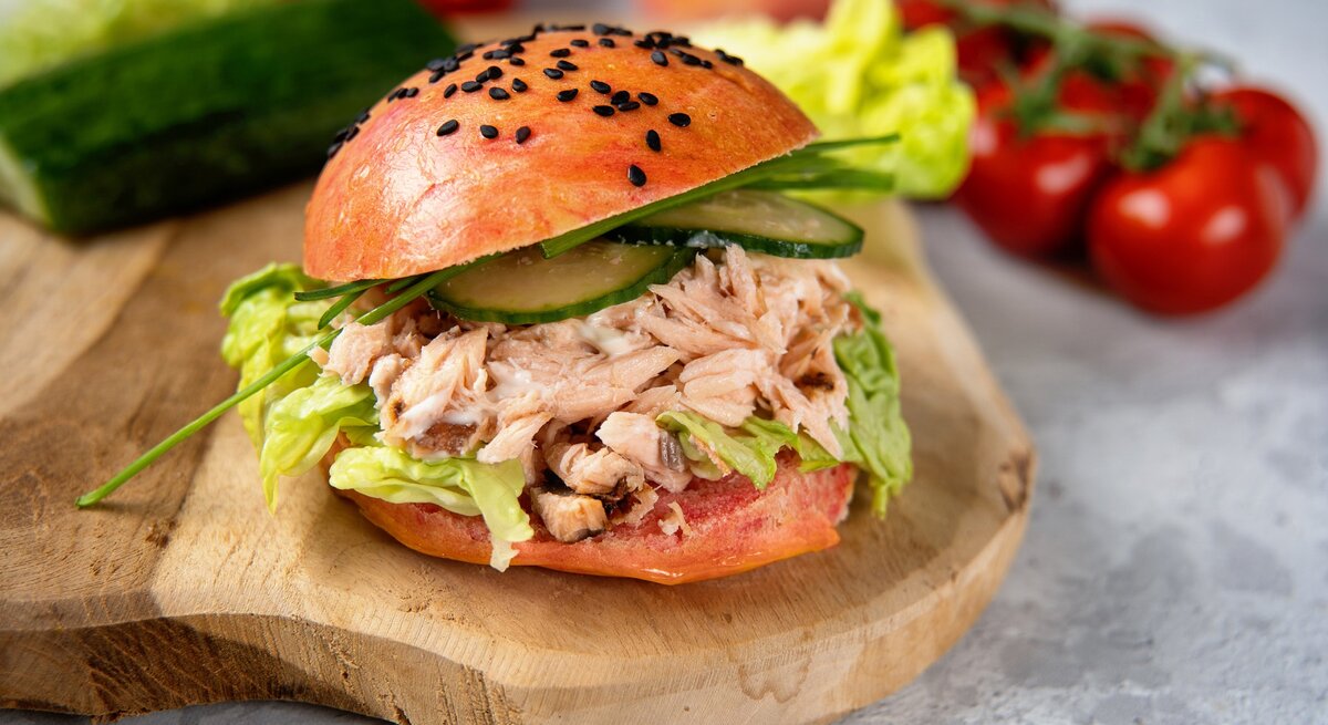 Pulled-Lachs-Burger auf einem Holzbrett. Im Hintergrund eine Gurke, Salat, Tomaten und Zwiebeln. 