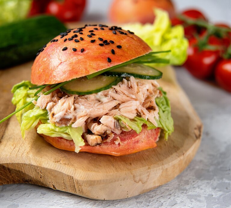 Pulled-Lachs-Burger auf einem Holzbrett. Im Hintergrund eine Gurke, Salat, Tomaten und Zwiebeln.  | © Getty Images/ Yummy pic