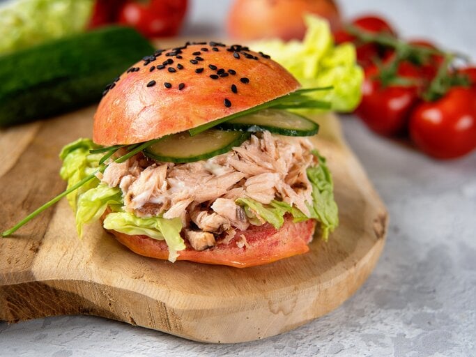 Pulled-Lachs-Burger auf einem Holzbrett. Im Hintergrund eine Gurke, Salat, Tomaten und Zwiebeln.  | © Getty Images/ Yummy pic