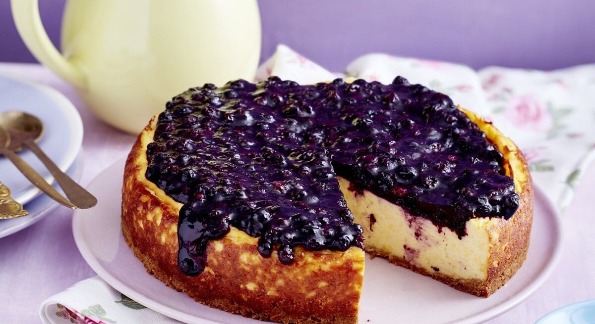 Auf einem Teller steht ein Heidelbeer-Pudding-Kuchen. Er ist sehr cremig und mit Heidelkompott bedeckt.
