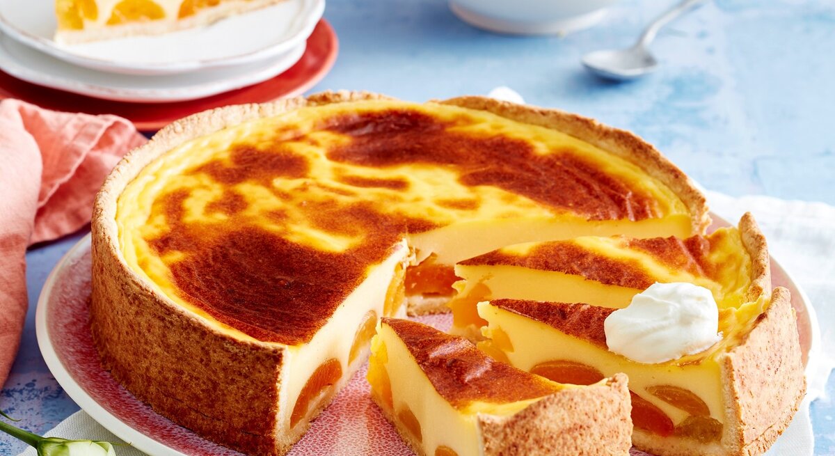 Angeschnittener Aprikosen-Puddingkuchen auf einem rötlichen Teller. Im Hintergrund ein einzelnes Stücj und eine Schüssel Sahne, alles auf blauem Untergrund. 