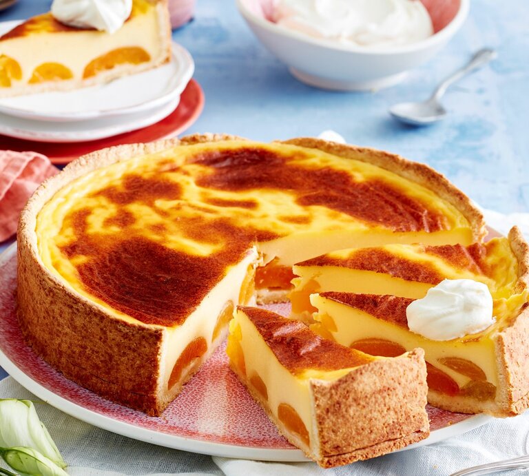 Angeschnittener Aprikosen-Puddingkuchen auf einem rötlichen Teller. Im Hintergrund ein einzelnes Stücj und eine Schüssel Sahne, alles auf blauem Untergrund.  | © EAT CLUB / Isabell Triemer