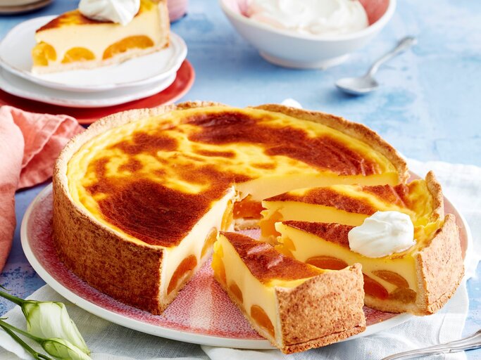 Angeschnittener Aprikosen-Puddingkuchen auf einem rötlichen Teller. Im Hintergrund ein einzelnes Stücj und eine Schüssel Sahne, alles auf blauem Untergrund.  | © EAT CLUB / Isabell Triemer