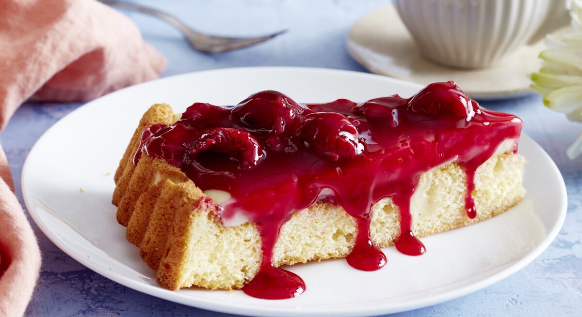 Pudding-Kirschkuchen auf einem weißen Teller, blauer Untergrund und Geschirr und Küchentuch im Hintergrund. 